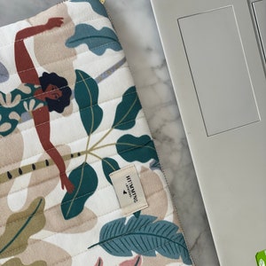 Peut inclure: Une housse d'ordinateur portable blanche avec un motif matelassé représentant une illustration colorée d'une personne dans un décor tropical. La housse est dotée d'un tirette de fermeture éclair en cuir marron et d'une petite étiquette portant la mention "HUMMING STUDIO".