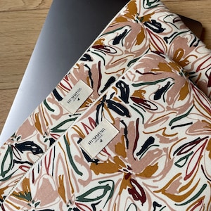 Puede incluir: Una funda para portátil blanca con un estampado floral abstracto de colores. La funda tiene una etiqueta marrón con el texto "HUMMING" y un pequeño logotipo de pájaro.