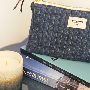 May include: A blue striped zippered pouch with a brown leather pull tab and a white label that reads "HUMMING atelier.". The pouch is sitting on top of two books. The top book is blue and has the text "LOUIS VUITTON CITY GUIDE AMSTERDAM" and "N PAVILIONS" on the cover. The bottom book is black and has the text "PHILIP JODIDIO" on the cover.