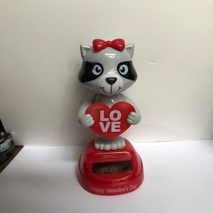 May include: A solar powered, bobbling raccoon figurine holding a red heart with the word "LOVE" on it. The base of the figurine is red and says "Happy Valentine's Day".