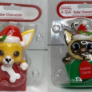 May include: Two solar powered Christmas figurines. A yellow dog wearing a Santa hat and holding a bone is in a red box. A brown cat wearing a Santa hat is in a green teacup with a red tag that says "Holiday Catnip".