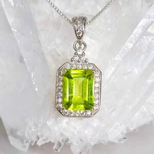 May include: A silver necklace featuring a rectangular, light green peridot pendant. The pendant is surrounded by a halo of small, clear gemstones. The necklace chain is delicate and silver. The pendant is approximately 0.75 inches long.