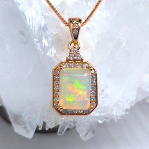 May include: A gold necklace with a square opal pendant surrounded by small clear stones. The pendant is on a white crystal background.