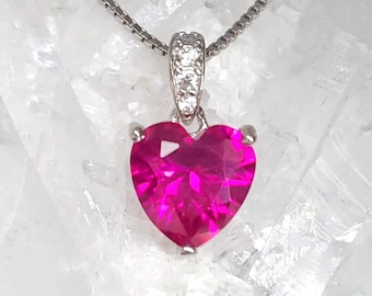 May include: A silver necklace with a heart-shaped, bright pink gemstone pendant. The pendant is set in a silver bezel and has a small, sparkling bail. The chain is a delicate silver chain.