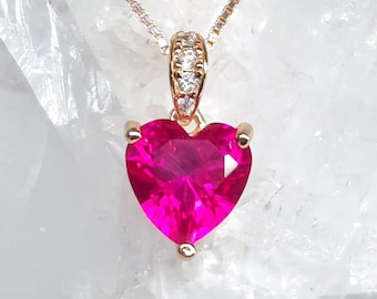May include: A gold-toned necklace featuring a heart-shaped, vibrant pink gemstone pendant. The pendant is suspended from a delicate chain and accented with small, clear gemstones. The necklace is displayed against a white, crystalline background.