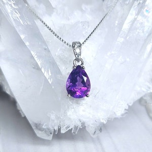 May include: A silver chain necklace with a pear-shaped amethyst pendant. The pendant is set in a silver bezel and has a faceted cut. The necklace is displayed on a white crystal background.