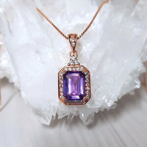 May include: A rose gold chain necklace with a pendant featuring a large, rectangular, purple amethyst gemstone surrounded by smaller, clear gemstones.