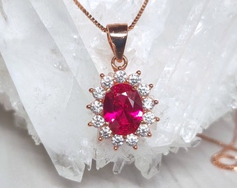 May include: A rose gold necklace with a pendant featuring a large oval-shaped ruby surrounded by a halo of smaller white cubic zirconia stones.