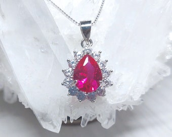May include: A silver pendant necklace with a teardrop-shaped red gemstone surrounded by a halo of smaller clear gemstones.