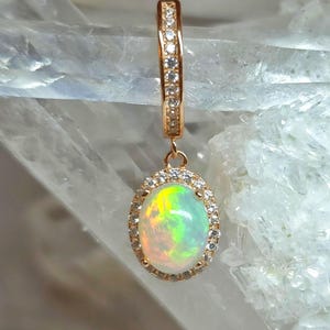 Könnte beinhalten: Goldfarbener Ohrring mit einem schimmernden, ovalen Opalanhänger. Der Opal ist von kleinen, klaren Steinen umgeben. Der obere Teil des Ohrrings ist ebenfalls mit kleinen, klaren Steinen besetzt.