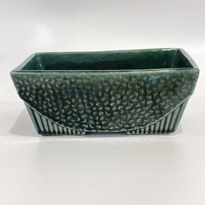 Pode incluir: Um vaso retangular de cerâmica com um esmalte verde-azulado profundo. O exterior apresenta um padrão texturizado de covinhas no painel central, enquanto as secções inferiores exibem estrias verticais. O recipiente tem uma abertura larga.