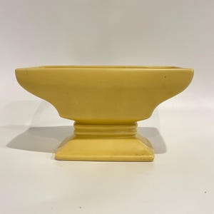 Puede incluir: Una maceta de cerámica de color amarillo mate con una parte superior rectangular ancha que se estrecha hacia un cuello central, apoyada sobre una base cuadrada escalonada. El recipiente presenta líneas suaves y un acabado amarillo uniforme.