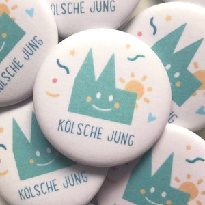Könnte beinhalten: Eine Sammlung weißer runder Knöpfe mit einem grünen Cartoon-Gebäude-Design. Das Gebäude hat ein lächelndes Gesicht und eine gelbe Sonne. Der Text "KÖLSCH JUNK" ist auf jedem Knopf gedruckt.