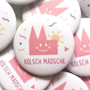 Könnte beinhalten: Ein Satz weißer runder Knöpfe mit einem rosa Comic-Haus mit einem lächelnden Gesicht und dem Text "KÖLSCH MÄDSCH" in rosa. Die Knöpfe haben einen bunten Hintergrund mit Sternen, Herzen und Wellenlinien.