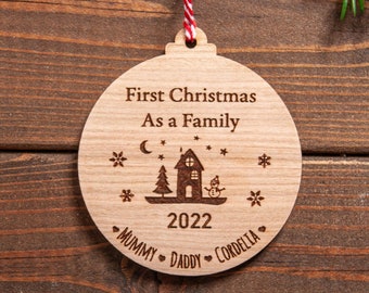 May include: A wooden Christmas ornament with the text "First Christmas As a Family" and the year "2022". The ornament also features a small house with a star above it, a moon, and snowflakes. The names "Mummy", "Daddy", and "Cordelia" are written below the house.