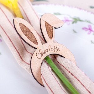 Op de afbeelding: Een houten bunny servetring met de naam "Charlotte" erop gegraveerd. De ring houdt een beige en rood gestreepte servet vast met een gele bloem.