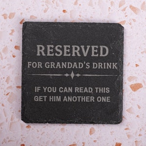 Op de afbeelding: Zwarte leisteen onderzetter met de tekst "RESERVED FOR GRANDAD'S DRINK. IF YOU CAN READ THIS GET HIM ANOTHER ONE."
