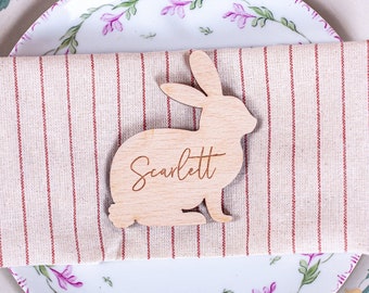 May include: A wooden bunny place card with the name "Scarlett" engraved on it. The place card is sitting on a striped linen napkin on a white plate with a floral design.