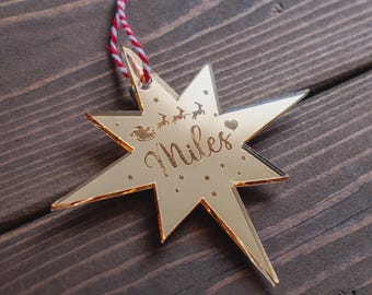 May include: A gold-colored star-shaped Christmas ornament with the name "Miles" engraved on it. The ornament features a red and white striped string for hanging and a small illustration of Santa's sleigh and reindeer.