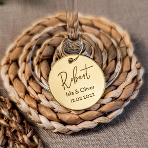 May include: A gold wine glass charm with the name "Robert", the names "Isla & Oliver" and the date "12.02.2022" engraved. The charm hangs from a wine glass, resting on a woven coaster made of natural fibres.