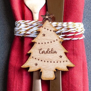 Puede incluir: Un adorno de árbol de Navidad de madera con el nombre "Cordelia" inscrito en él. El adorno está atado a una servilleta roja con cuerda blanca.