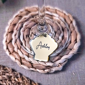 Puede incluir: Un posavasos tejido con una copa de vino y un charm en forma de concha. El charm es blanco con el nombre "Ashley" escrito en cursiva. El posavasos está hecho de fibras naturales y es marrón claro y beige.