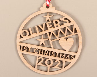 May include: A wooden Christmas ornament with a cutout design of a Christmas tree, a heart, and stars. The ornament is personalized with the text "Oliver's 1st Christmas 2021".