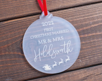 Pode incluir: Uma bola de Natal de vidro fosco com uma fita vermelha. O enfeite diz "2022 First Christmas Married Mr & Mrs Holdsworth" com um desenho de Papai Noel e renas. Em um fundo de madeira.