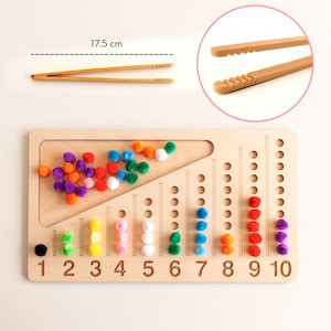 Puede incluir: Tablero de conteo de madera con pompones de colores y números del 1 al 10. El tablero mide 17,5 cm de largo e incluye un par de pinzas de madera.