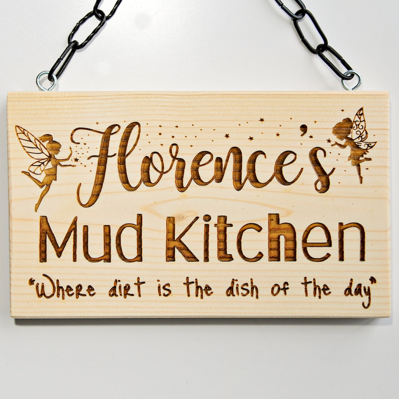 Peut inclure: Une pancarte en bois avec un motif de f&eacute;e et le texte "Florence's Mud Kitchen" et la citation "Where dirt is the dish of the day".