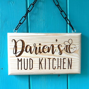 Peut inclure: Une pancarte en bois avec une cha&icirc;ne pour l'accrocher, indiquant "Darien's Mud Kitchen" avec une illustration de dessin anim&eacute; d'une abeille.