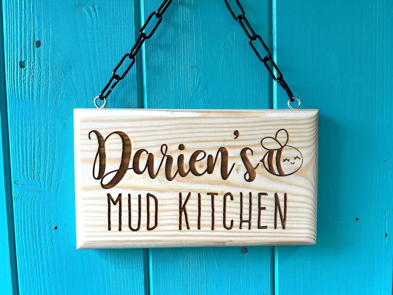 Peut inclure: Une pancarte en bois avec une cha&icirc;ne pour l'accrocher, indiquant "Darien's Mud Kitchen" avec une illustration de dessin anim&eacute; d'une abeille.