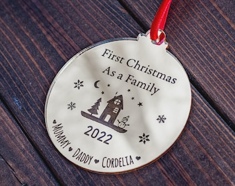 May include: A round Christmas ornament with the text "First Christmas As a Family" and the year 2022. The ornament features a house, trees, snowflakes, and a snowman design. It includes the names "Mummy, Daddy, Cordelia" and hangs from a red ribbon.