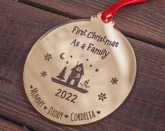 May include: A gold-colored Christmas ornament with a red ribbon. The ornament has the text "First Christmas As a Family" and the year "2022". It features a house, a snowman, a tree, stars, and the names "Mummy", "Daddy", and "Cordelia".