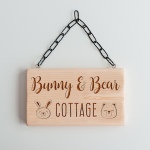 Peut inclure: Une pancarte en bois avec une cha&icirc;ne noire suspendue. La pancarte indique "Bunny & Bear Cottage" avec une illustration de lapin et d'ours de chaque c&ocirc;t&eacute;.