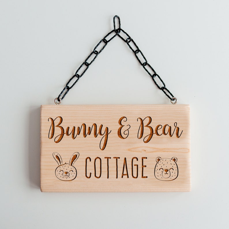 Peut inclure: Une pancarte en bois avec une cha&icirc;ne noire suspendue. La pancarte indique "Bunny & Bear Cottage" avec une illustration de lapin et d'ours de chaque c&ocirc;t&eacute;.
