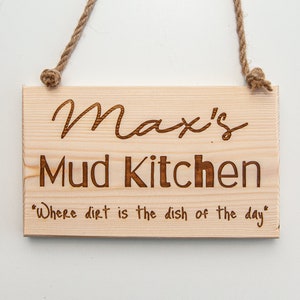 Peut inclure: Une pancarte en bois avec le texte "Max's Mud Kitchen" et la citation "Where dirt is the dish of the day". La pancarte est suspendue &agrave; une corde.