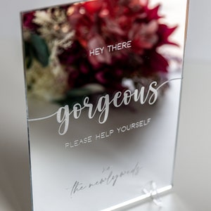 May include: A silver mirror sign with the text "Hey there gorgeous, please help yourself to the newlyweds".