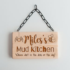 Peut inclure: Une pancarte en bois avec le texte "Miles's Mud Kitchen" et la phrase "Where dirt is the dish of the day". La pancarte est d&eacute;cor&eacute;e d'une araign&eacute;e et d'une abeille.