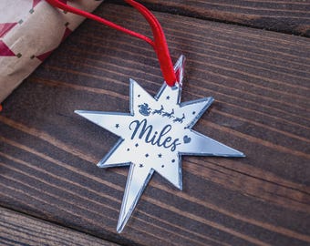 May include: A mirrored star-shaped Christmas ornament with the name "Miles" in script. The ornament features a silhouette of Santa and reindeer, with a red ribbon for hanging. The background is a dark wooden surface.