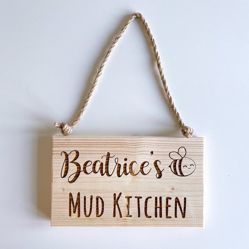 Peut inclure: Une pancarte en bois avec un cintre en corde, indiquant "Beatrice's Mud Kitchen" avec une illustration de dessin anim&eacute; d'une abeille.