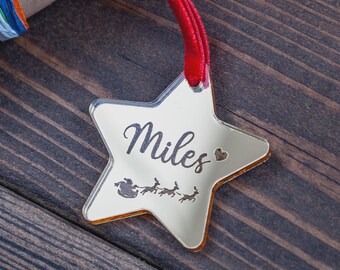 May include: A star-shaped ornament with the name "Miles" engraved in a cursive font. The ornament has a red ribbon for hanging and features a silhouette of Santa's sleigh and reindeer. The ornament is made of a reflective material.
