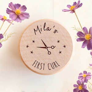 May include: A round, light-colored wooden keepsake box lid featuring the engraved text 'Mila's FIRST CURL' surrounding a central graphic of a pair of scissors flanked by four small stars, with purple cosmos flowers visible in the corners.
