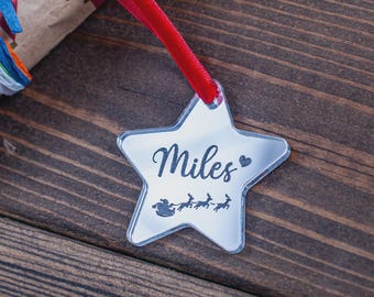 May include: A star-shaped Christmas ornament with the name "Miles" in cursive script, a heart, and a silhouette of Santa's sleigh and reindeer. The ornament hangs from a red ribbon. The background is a wooden surface.