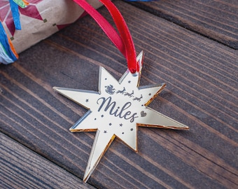 May include: A gold star-shaped Christmas ornament with the name "Miles" in script. The ornament has a red ribbon for hanging and features a silhouette of Santa and reindeer. The ornament is on a dark wood surface.