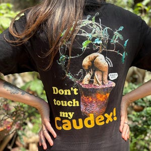 Puede incluir: Camiseta negra con un gráfico de una planta caudex en una maceta. El texto "Don't touch my caudex!" está impreso en amarillo en la camiseta.