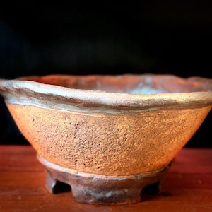Puede incluir: Un tiesto de bonsái de cerámica hecho a mano con una superficie texturizada. El tiesto presenta una paleta de colores cálidos y terrosos, con un borde claro y una base oscura. El tiesto en forma de cuenco está diseñado para bonsáis y otras plantas pequeñas.