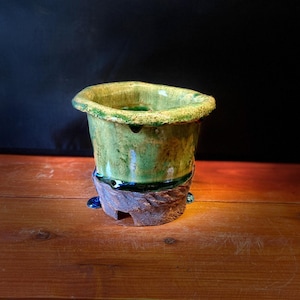 May include: A handmade ceramic vase with a unique, organic shape. The vase features a vibrant green glaze with a textured rim and a dark brown base. The base has a rough, earthy texture, and the vase is set on a wooden surface.