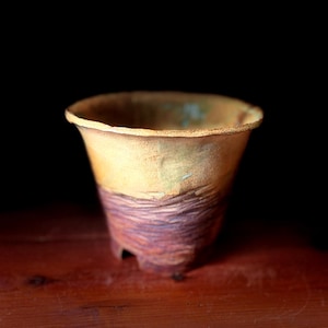 May include: A handmade ceramic cup with a textured, layered design. The cup features a light yellow upper portion and a lower section with purple and brown tones. The cup has a flared rim and sits on three small feet.