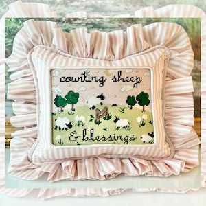 May include: A decorative pillow with a cross-stitched design featuring sheep in a field, trees, and the text "counting sheep & blessings." The pillow has a pink and white striped border and a ruffled edge.